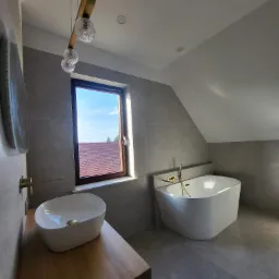 Nowoczesna łazienka na poddaszu z wolnostojącą wanną, umywalką nablatową na drewnianym blacie i szarą glazurą, oświetlona designerską lampą z trzema szklanymi kulami.