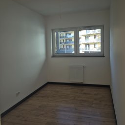 Fenix Grzegorz Woźniak - Pusty pokój po remoncie: drewnopodobne panele, białe ściany, grzejnik pod oknem z widokiem na blok. Nowe wykończenie wnętrza, gotowe do umeblowania.
