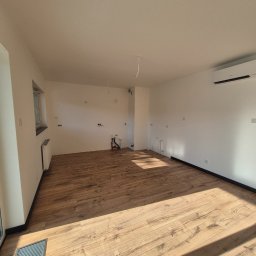 Fenix Grzegorz Woźniak - Jasne panele podłogowe w pustym pokoju z białymi ścianami i grzejnikiem. Widoczne instalacje wodne i klimatyzacja. Minimalistyczne wykończenie wnętrza.