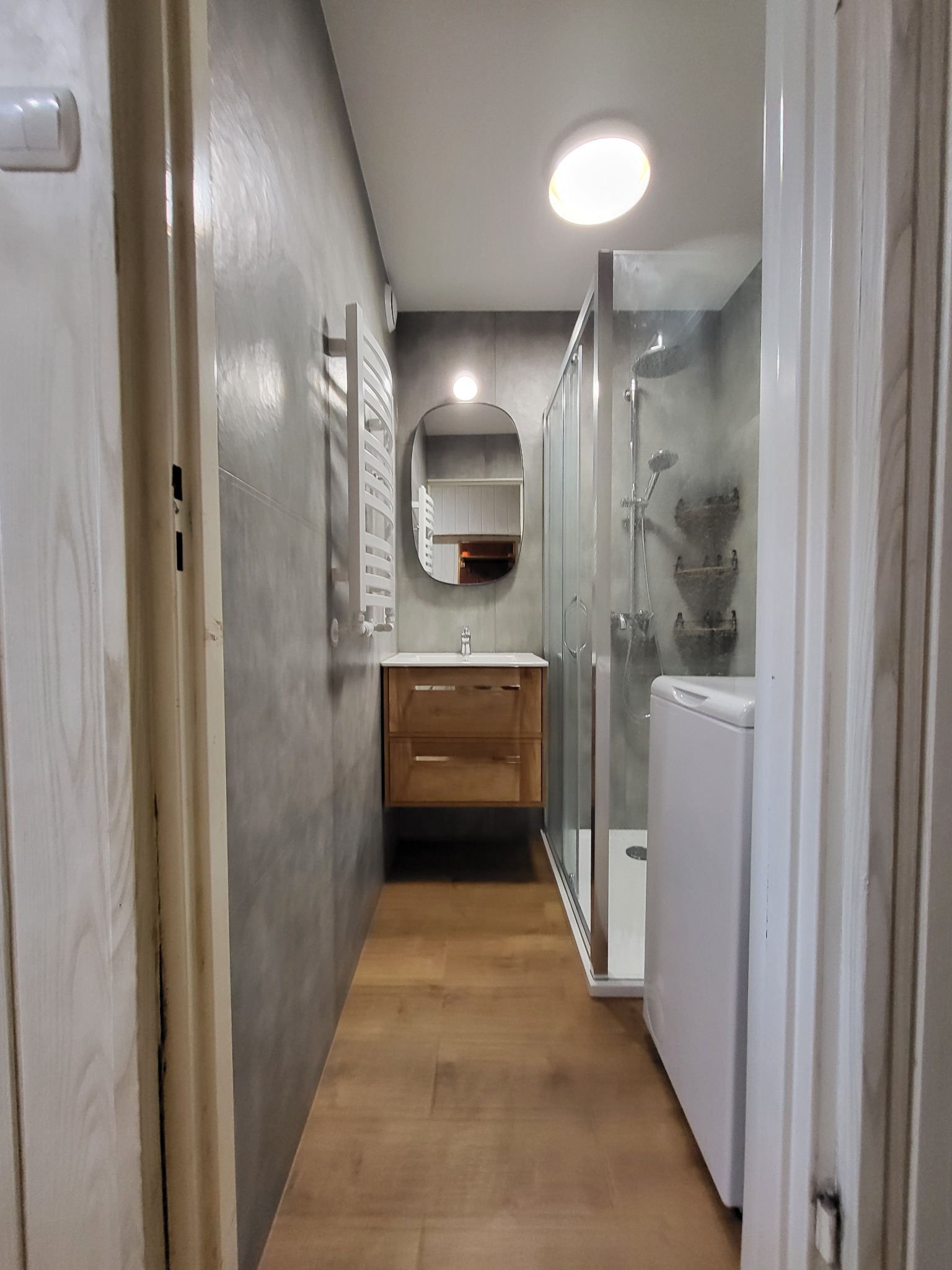 Wnętrze łazienki z szarą ścianą, drewnianą szafką pod umywalką, owalnym lustrem i kabiną prysznicową z pralką, widoczne zza uchylonych drzwi.