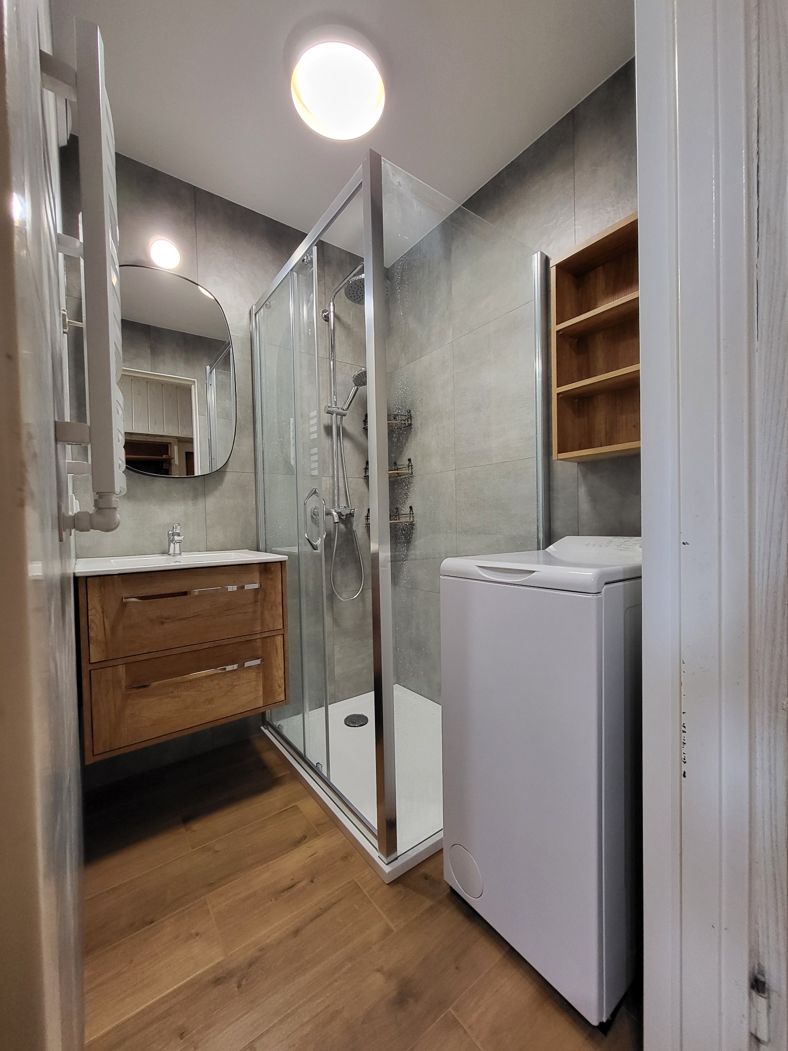 Wykończona łazienka z kabiną prysznicową, drewnianą szafką pod umywalką, grzejnikiem drabinkowym, drewnianą półką i pralką, szare płytki na ścianach i drewnopodobne na podłodze.
