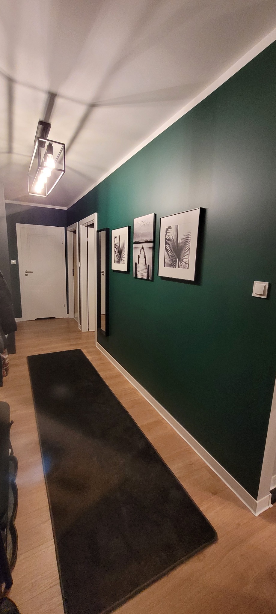 Nowoczesny przedpokój z ciemnozieloną ścianą, trzema czarno-białymi obrazami w ramkach, długim, ciemnym dywanem i drewnopodobną podłogą, oświetlony lampą sufitową z żarówkami Edisona.