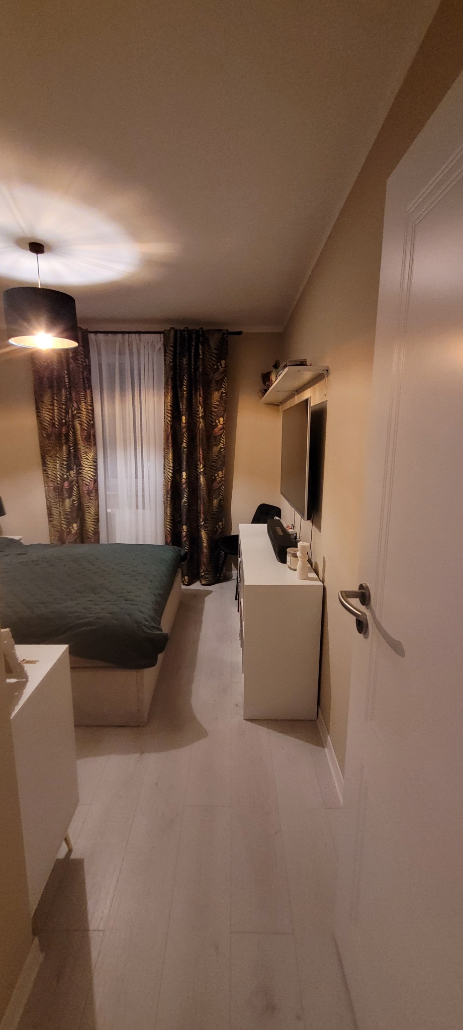 Sypialnia z łóżkiem z zieloną narzutą, zasłonami w tropikalne wzory, białymi meblami i telewizorem na ścianie, oświetlona lampą wiszącą z czarnym abażurem.