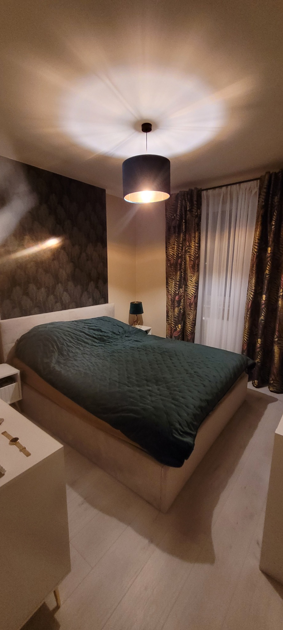 Nowoczesna sypialnia z łóżkiem kontynentalnym, ciemnozieloną narzutą i lampą sufitową z abażurem, widoczne tapety w motywy roślinne i zasłony w tropikalne wzory.