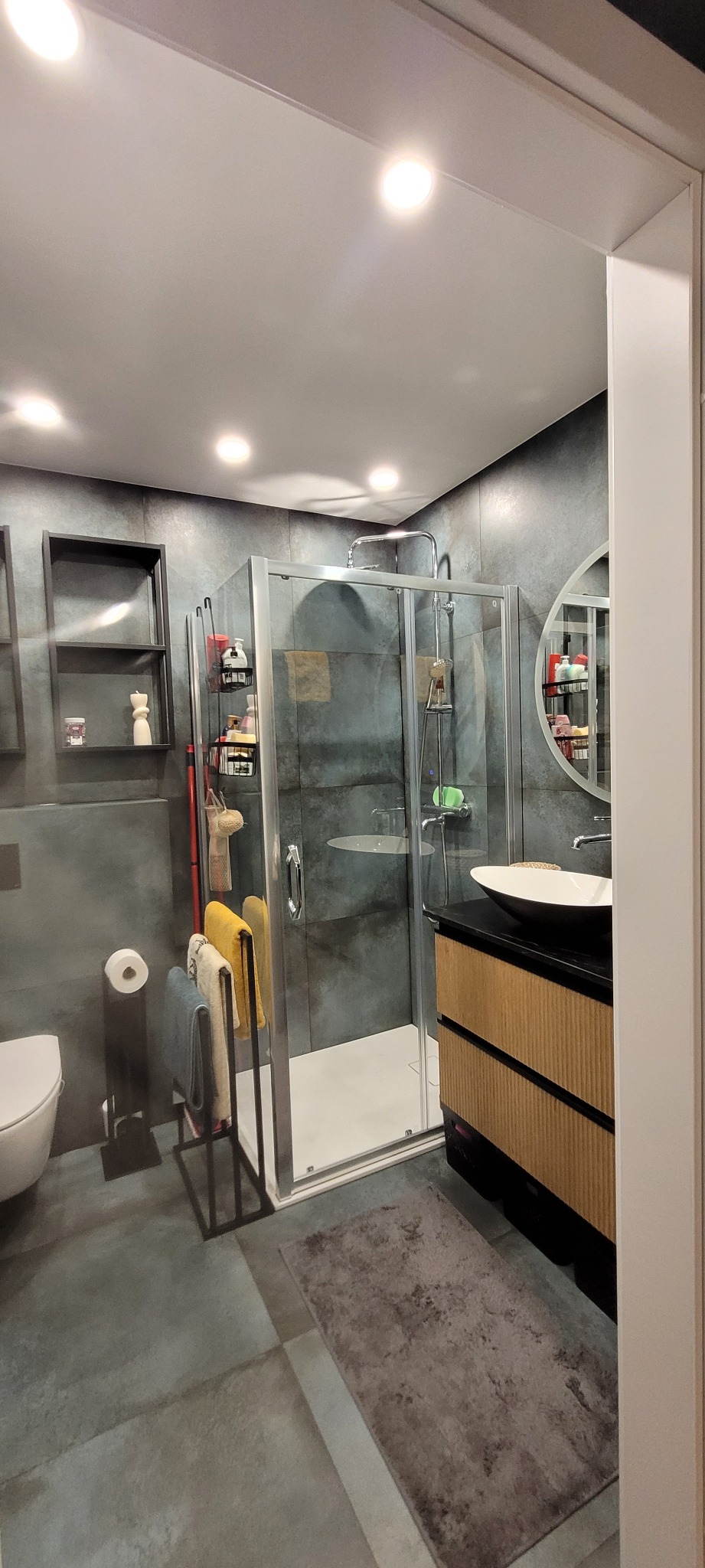 Nowoczesna łazienka z szarą płytką, kabiną prysznicową ze szklanymi drzwiami, umywalką nablatową na szafce z lamelami i okrągłym lustrem.