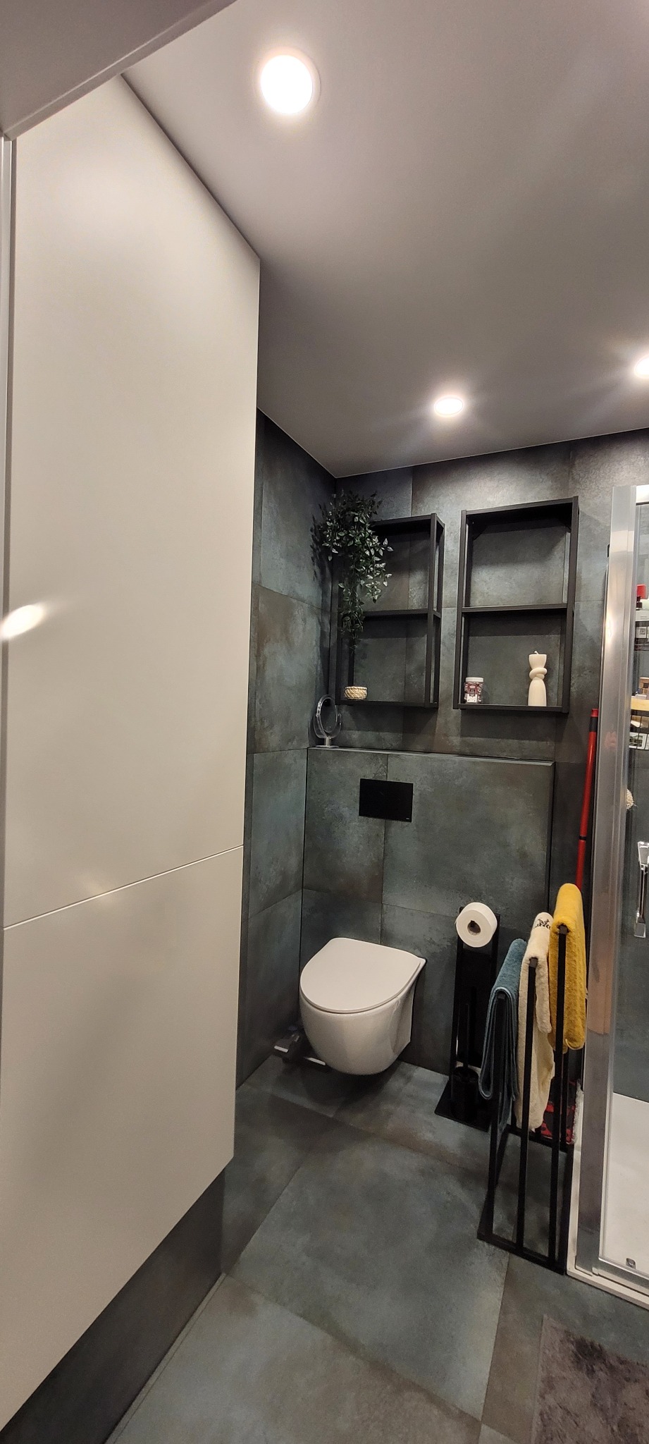 Nowoczesna łazienka z podwieszaną toaletą, grafitowymi płytkami, czarnymi półkami dekoracyjnymi z rośliną i ozdobami, oraz minimalistycznym grzejnikiem na ręczniki.