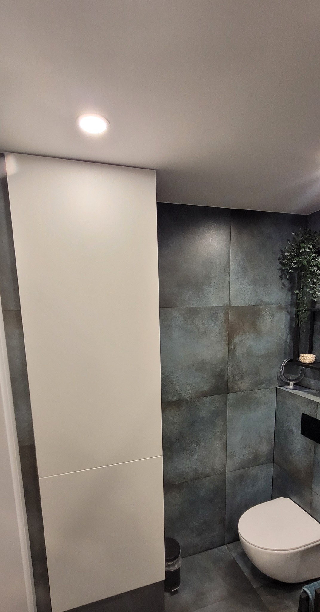 Nowoczesna łazienka we Wrocławiu z szarymi płytkami imitującymi beton, wisząca miska WC, biała zabudowa szafkowa i punktowe oświetlenie sufitowe.