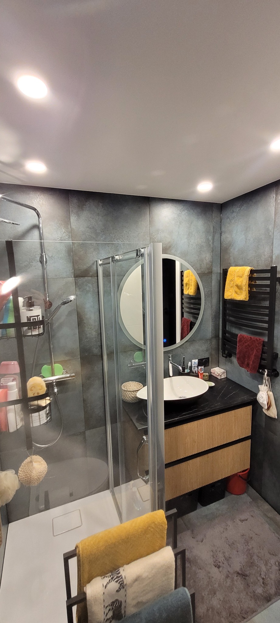 Nowoczesna łazienka z szarymi płytkami imitującymi beton, kabiną prysznicową z transparentnego szkła, okrągłym lustrem z podświetleniem LED, umywalką nablatową na szafce z drewnianymi frontami...