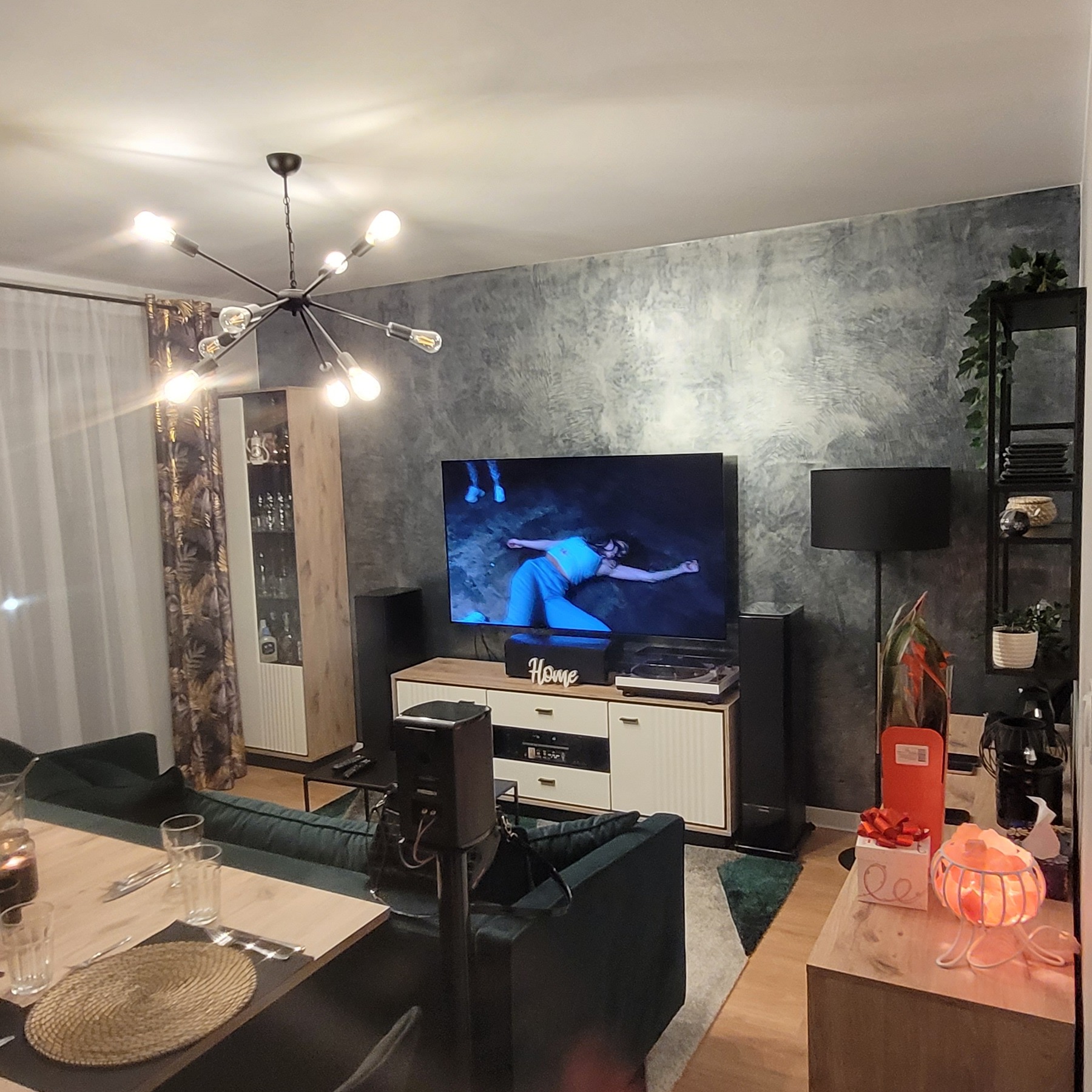 Nowoczesny salon z szarą ścianą o fakturze betonu, zieloną sofą, drewnianym stołem z nakryciem, designerską lampą sufitową i szafką RTV z gramofonem.