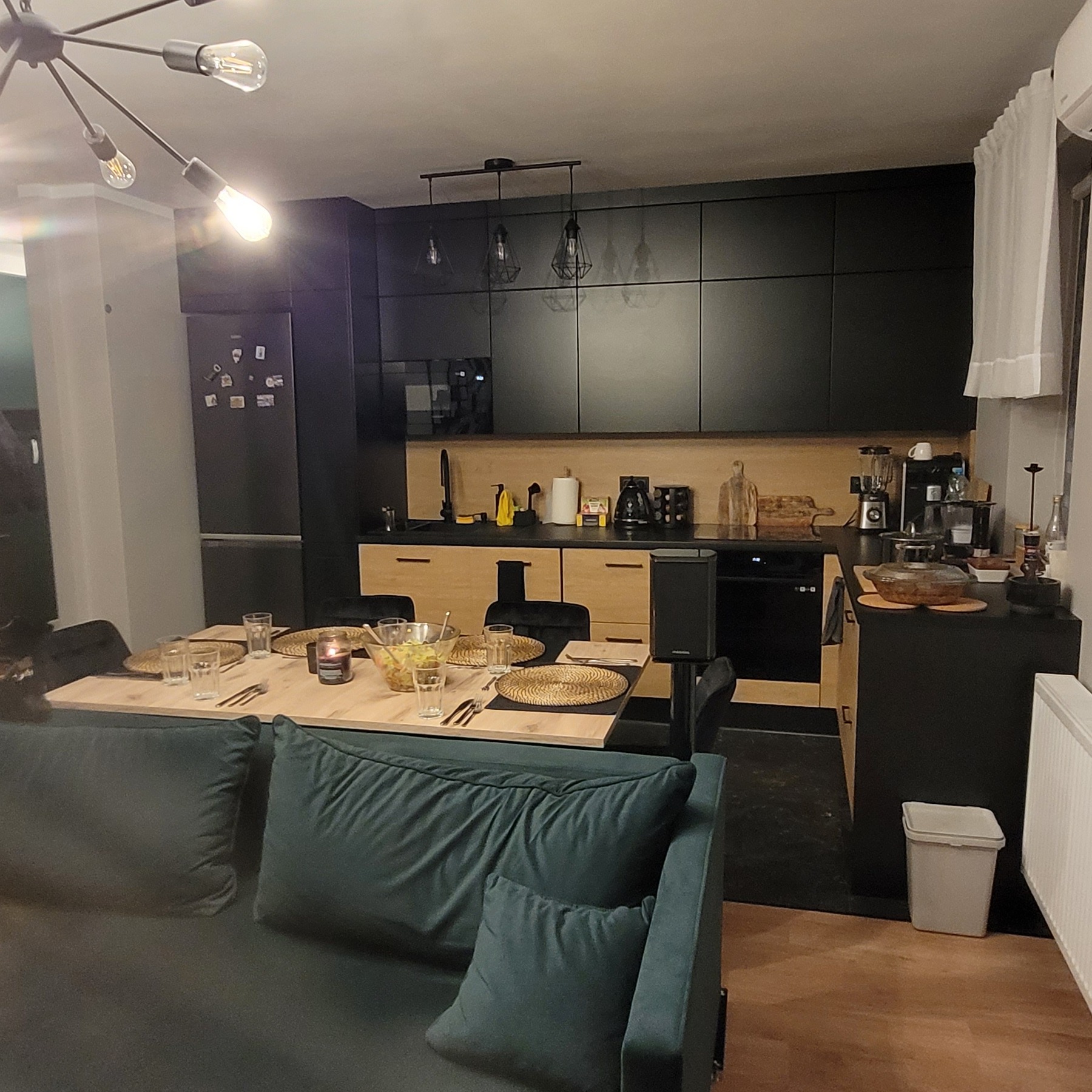 Nowoczesna kuchnia z czarnymi szafkami i drewnianymi akcentami, stół nakryty do posiłku z dekoracyjną świecą, widok z perspektywy salonu z zieloną sofą.