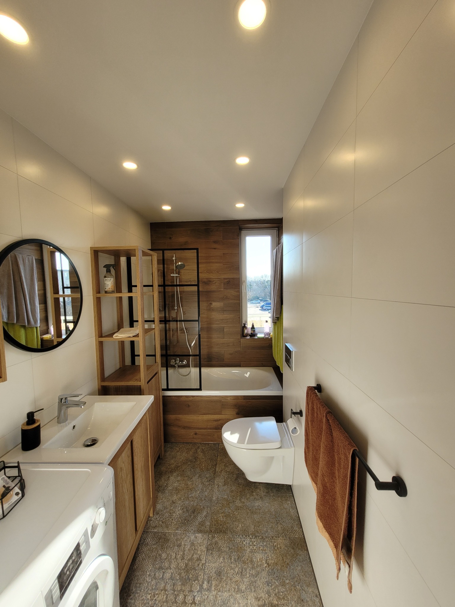 Nowoczesna łazienka z drewnianymi akcentami: płytki imitujące drewno, kabina prysznicowa typu walk-in z czarną ramą, umywalka z szafką z drewna, okrągłe lustro i biała pralka.