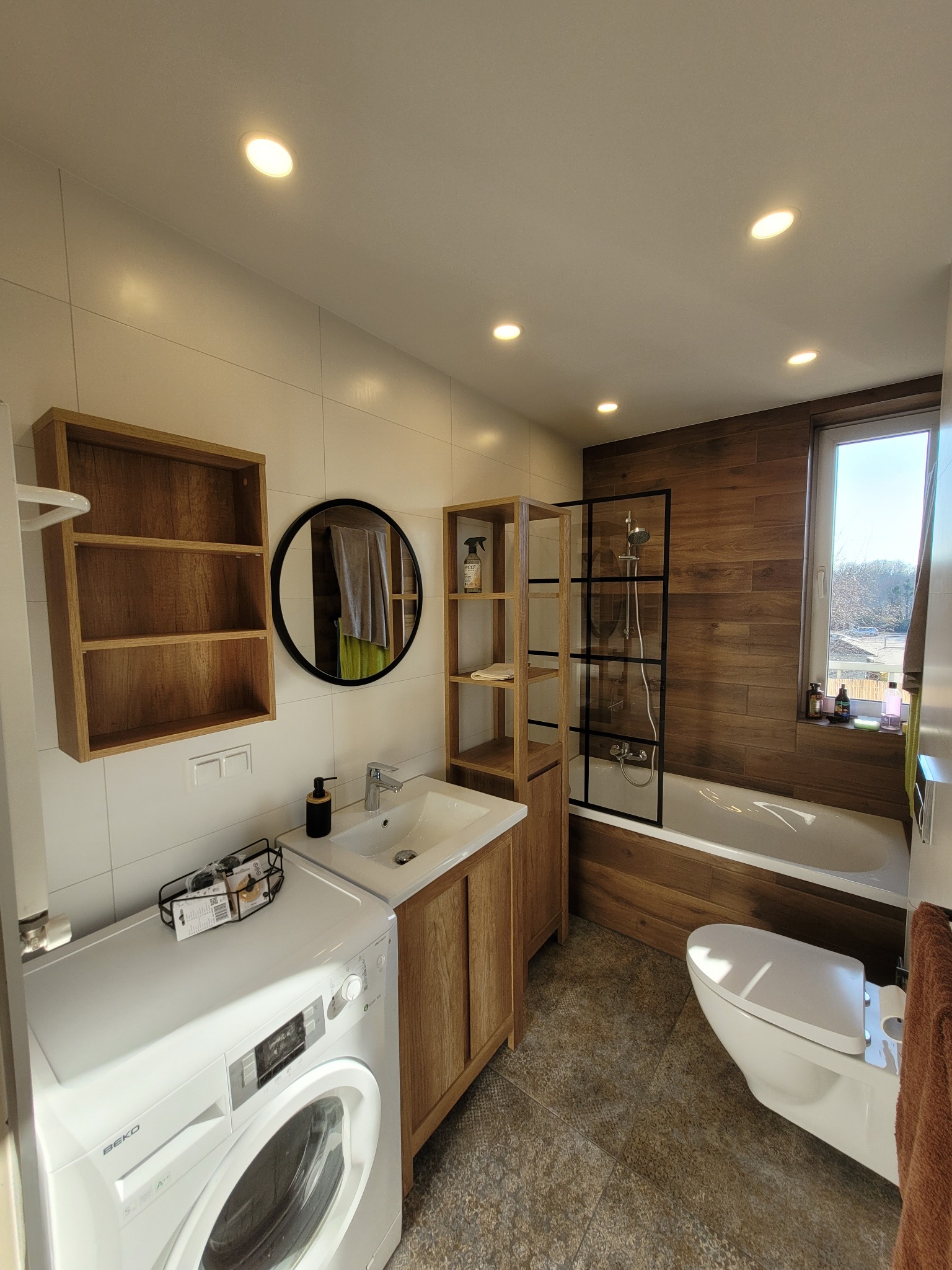 Nowoczesna łazienka z drewnianymi akcentami: szafki, półki, obudowa wanny i pralka Beko z koszykiem na akcesoria na górze.