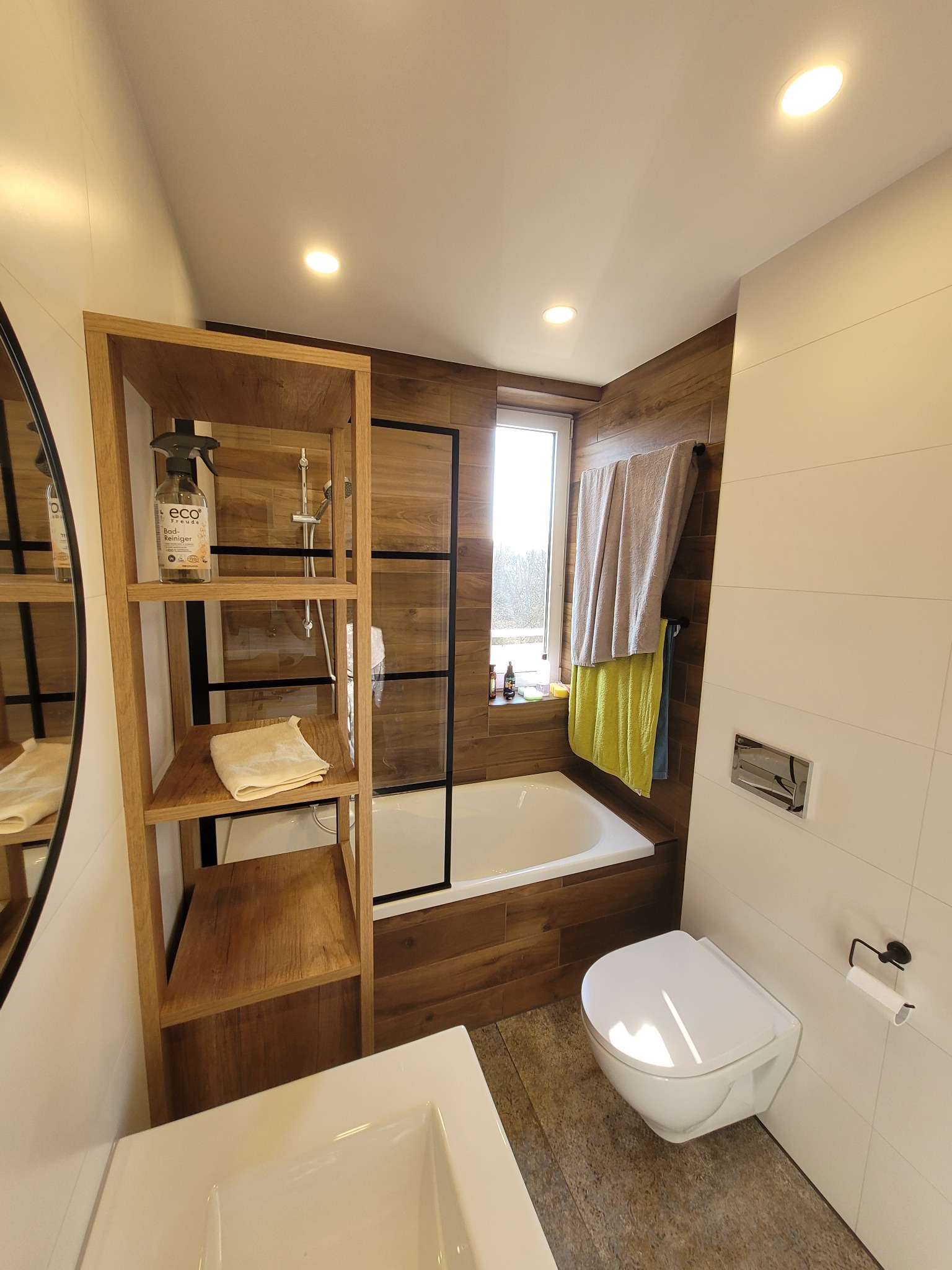 Nowoczesna łazienka z drewnianymi akcentami: półka z kosmetykami Eco Freude, wanna za czarną, szklaną ścianką, wisząca toaleta i ręczniki przy oknie.