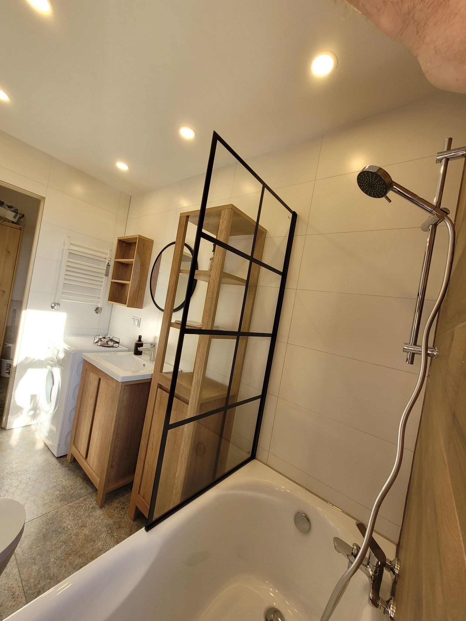 Nowoczesna łazienka z wanną, szklaną ścianką działową z czarnymi ramami, drewnianą szafką pod umywalką i półką, pralką oraz grzejnikiem drabinkowym.