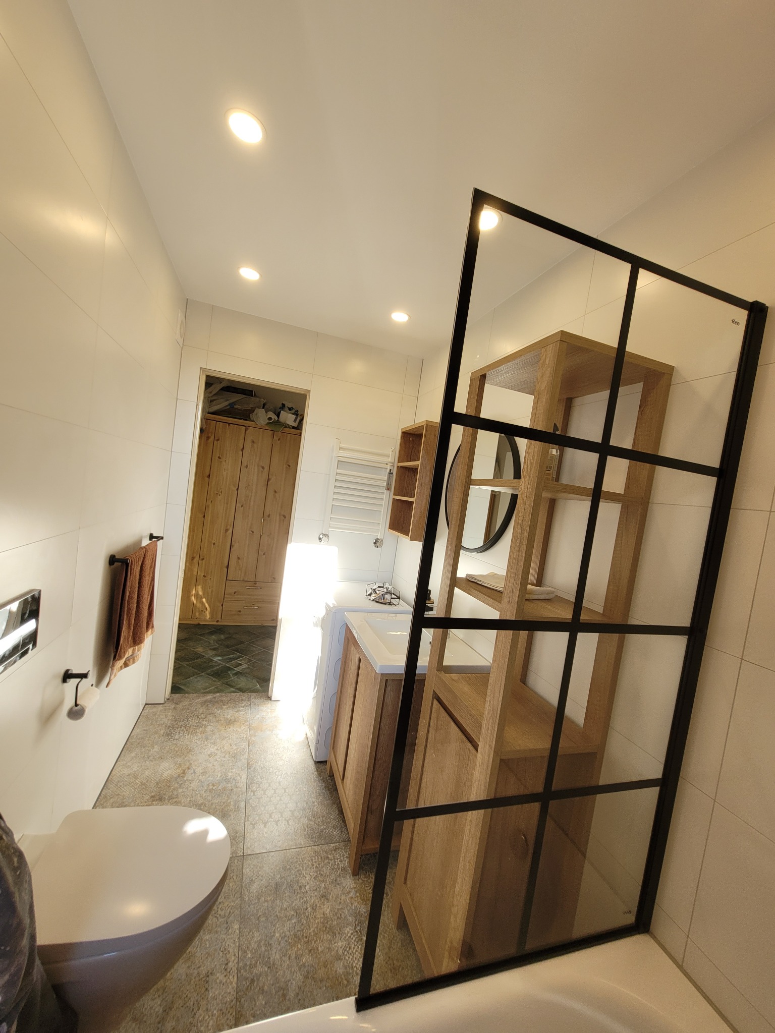 Jasna łazienka z drewnianą szafką pod umywalką, kabiną prysznicową z czarnymi szprosami i widoczną toaletą z przodu, wzorzysta podłoga, drzwi do garderoby w tle.