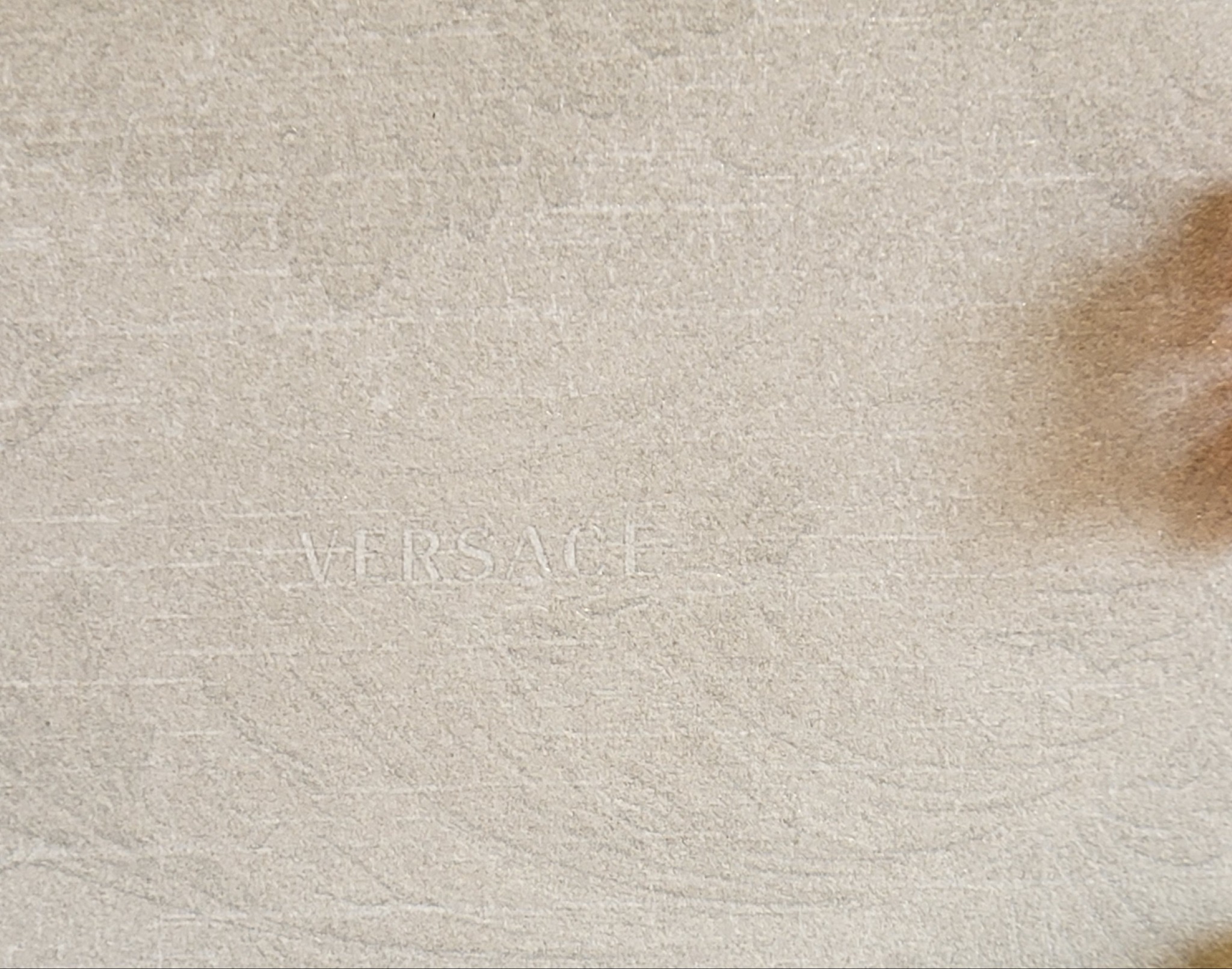 Beżowa płytka ceramiczna z subtelnym, wytłoczonym napisem 'VERSACE'.