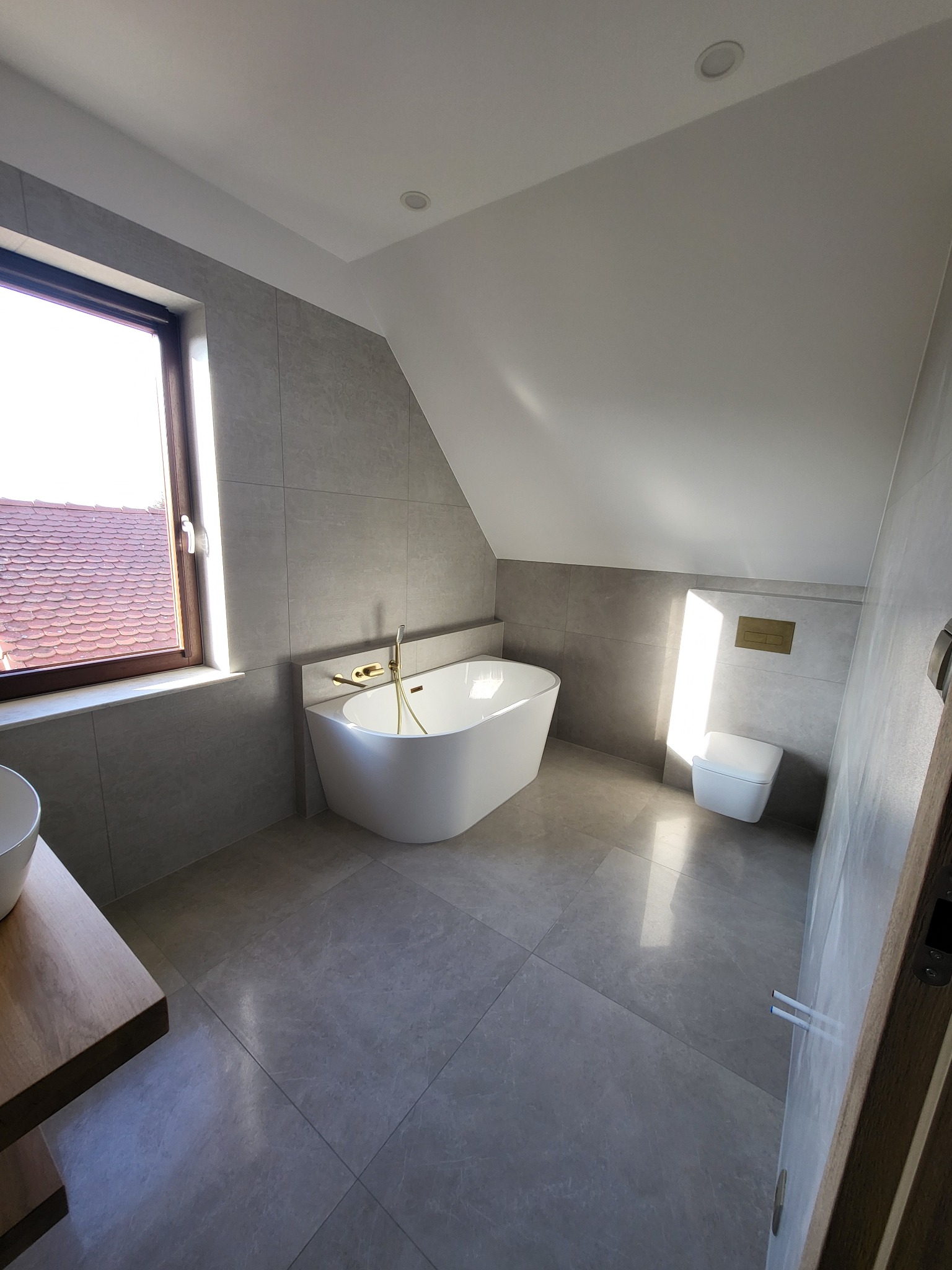 Jasna łazienka na poddaszu z wolnostojącą wanną, szarą glazurą wielkoformatową, drewnianym blatem umywalki i oknem dachowym z widokiem na czerwony dach.