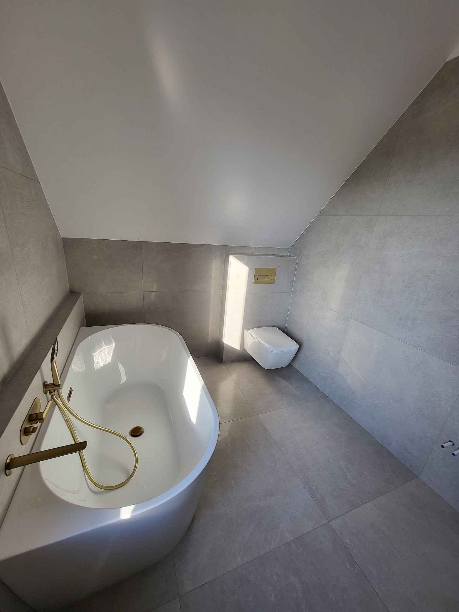 Nowoczesna łazienka na poddaszu z wanną wolnostojącą, złotą armaturą i podwieszaną toaletą, wykończona szarymi płytkami wielkoformatowymi.