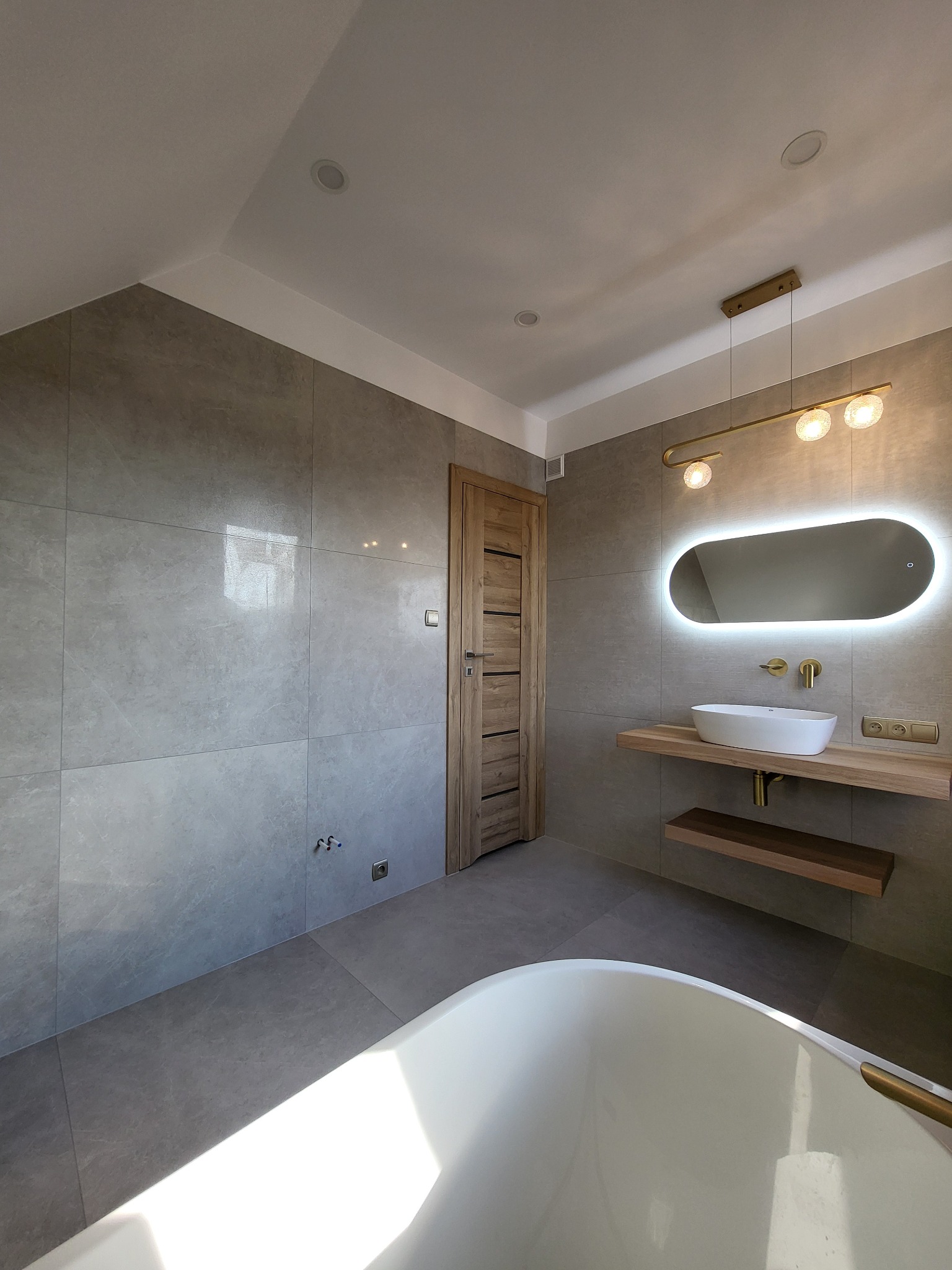 Nowoczesna łazienka z szarymi płytkami, podświetlanym owalnym lustrem nad umywalką na drewnianym blacie, złotą armaturą i lampą, oraz fragmentem białej wanny.