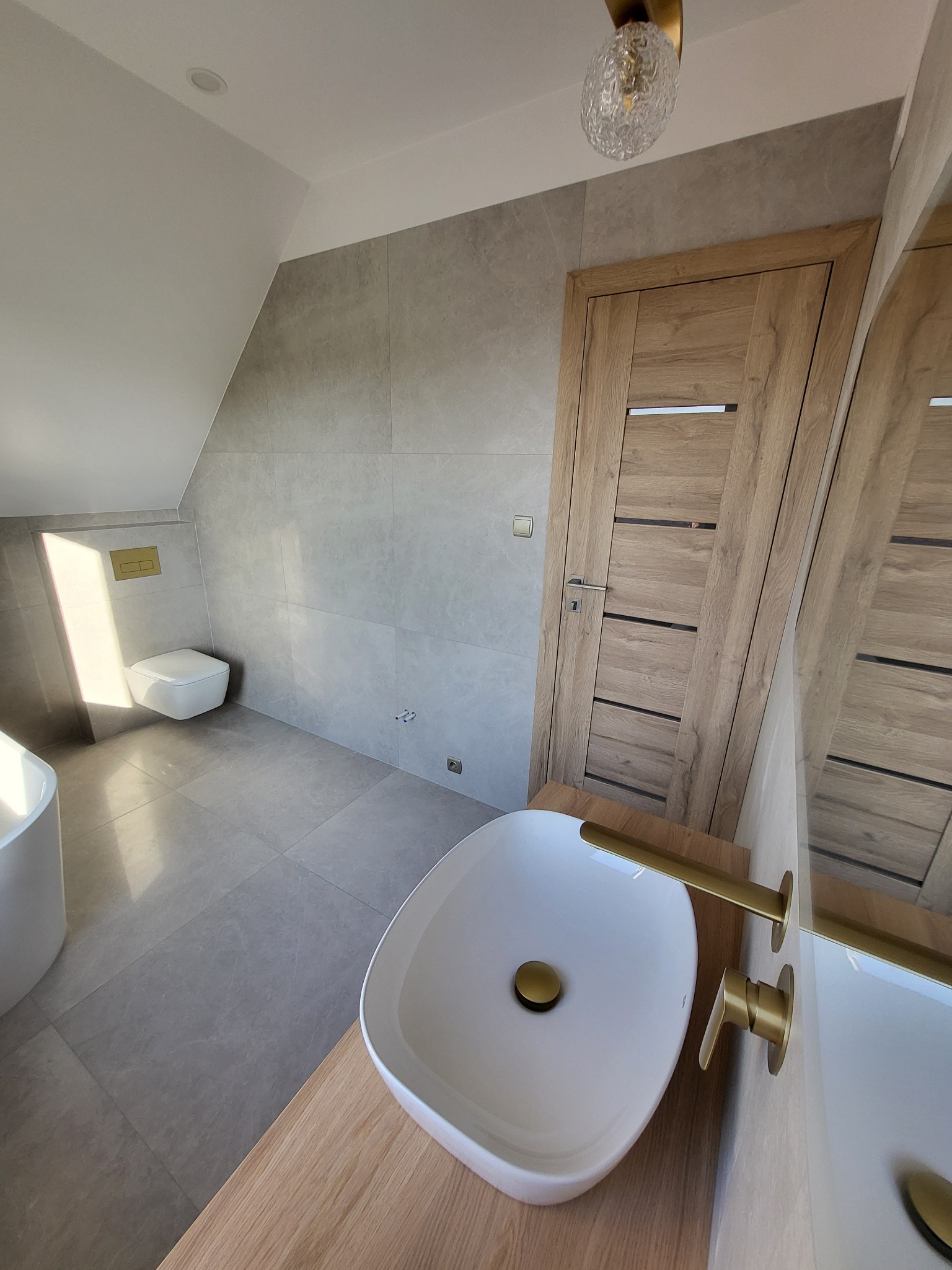 Nowoczesna łazienka z umywalką nablatową, baterią w kolorze złotym, szarą glazurą, drewnianymi drzwiami i wiszącą toaletą.