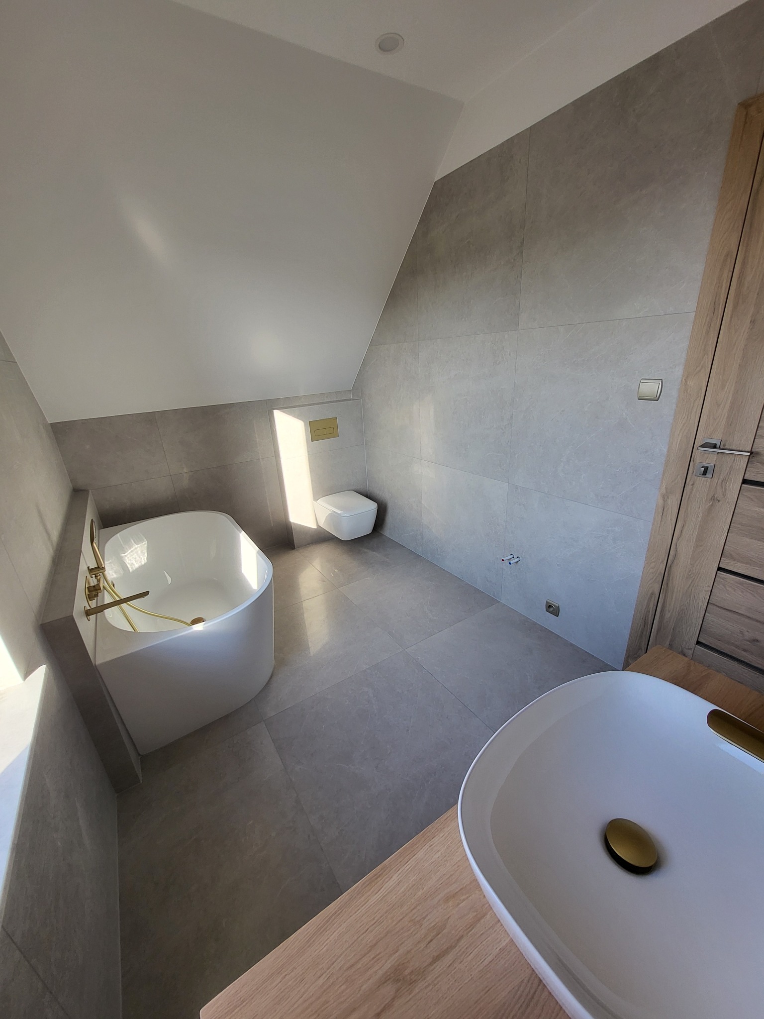 Nowoczesna łazienka na poddaszu z wanną wolnostojącą, umywalką nablatową z złotymi akcentami i szarą płytką wielkoformatową na ścianach.