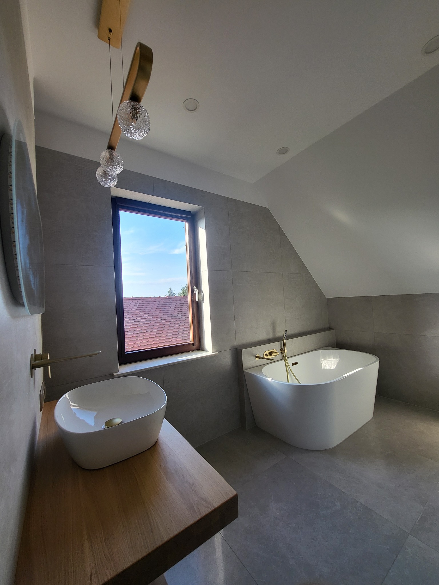 Nowoczesna łazienka na poddaszu z wolnostojącą wanną, umywalką nablatową na drewnianym blacie i szarą glazurą, oświetlona designerską lampą z trzema szklanymi kulami.