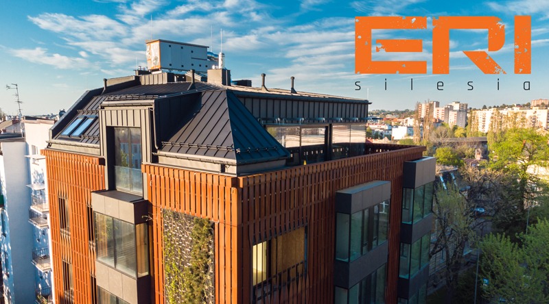 Nowoczesny budynek wielorodzinny z ciemnym, metalowym dachem, drewnianą elewacją i zieloną ścianą, z logotypem 'ERI Silesia' w prawym górnym rogu, widok z góry na tle panoramy miasta.
