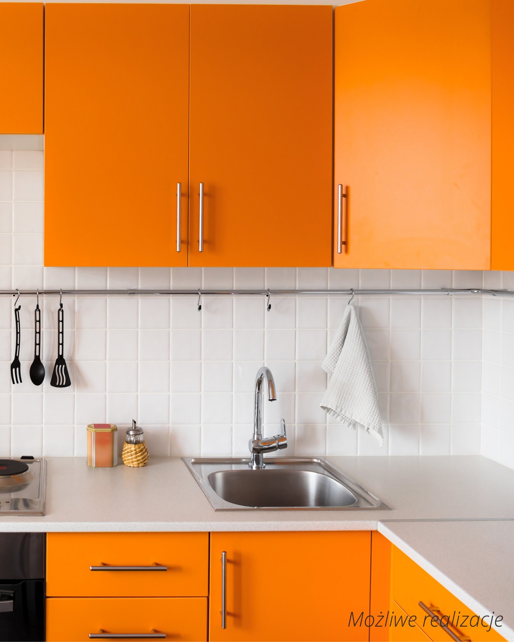 Fragment kuchni z pomarańczowymi szafkami, srebrnym zlewem i białymi płytkami na ścianie, z widocznymi akcesoriami kuchennymi zawieszonymi na relingu.