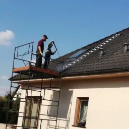 Dwóch pracowników na rusztowaniu czyści dach domu z użyciem myjki ciśnieniowej; widoczne okno dachowe i drabinka dachowa.