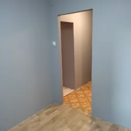 Wnętrze pokoju z pomalowanymi na szaro ścianami i panelami podłogowymi, widok na sąsiednie pomieszczenie z wzorzystą podłogą.