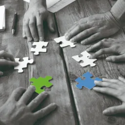 Zespół ludzi układa białe puzzle na drewnianym stole, z wyróżnionymi zielonym i niebieskim elementem, symbolizując współpracę i budowanie całości.