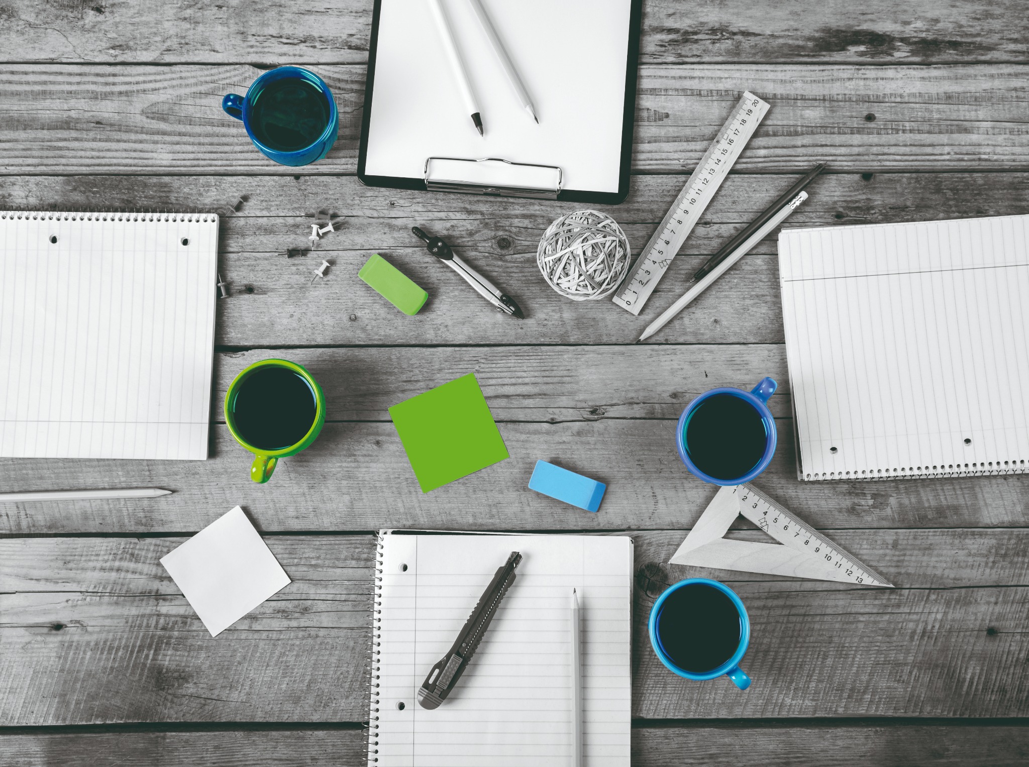 Widok z góry na biurko z drewnianym blatem, na którym znajdują się notatniki, długopisy, ołówki, gumki, linijka, cyrkiel, spinacze, karteczki samoprzylepne i cztery kubki z ciemnym napojem.