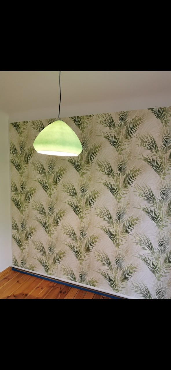 Ściana pokryta tapetą w motywy palmowe, oświetlona zieloną lampą wiszącą, z widoczną drewnianą podłogą.