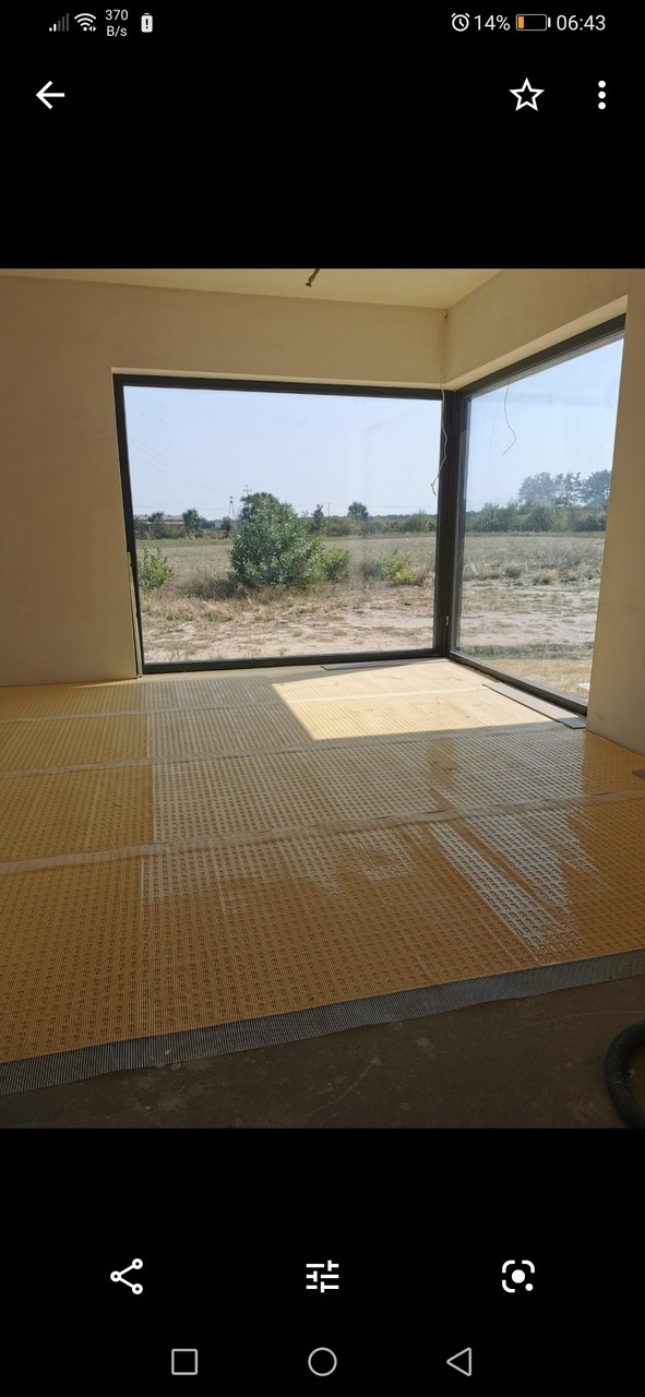 Przygotowanie podłoża pod płytki: żółta mata drenażowa ułożona na podłodze w surowym pomieszczeniu z dużym oknem z widokiem na łąkę.