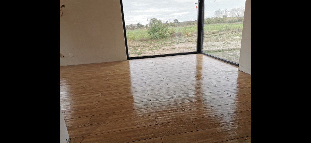 Ułożona, błyszcząca podłoga z płytek imitujących drewno w nowym pomieszczeniu z dużym oknem na zewnątrz, widoczne niedoskonałości na ścianie.