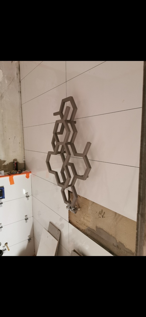 Montaż dekoracyjnego grzejnika w kształcie heksagonów na białej ścianie z płytek w łazience w trakcie remontu, widoczne niedokończone fragmenty ściany i narzędzia.