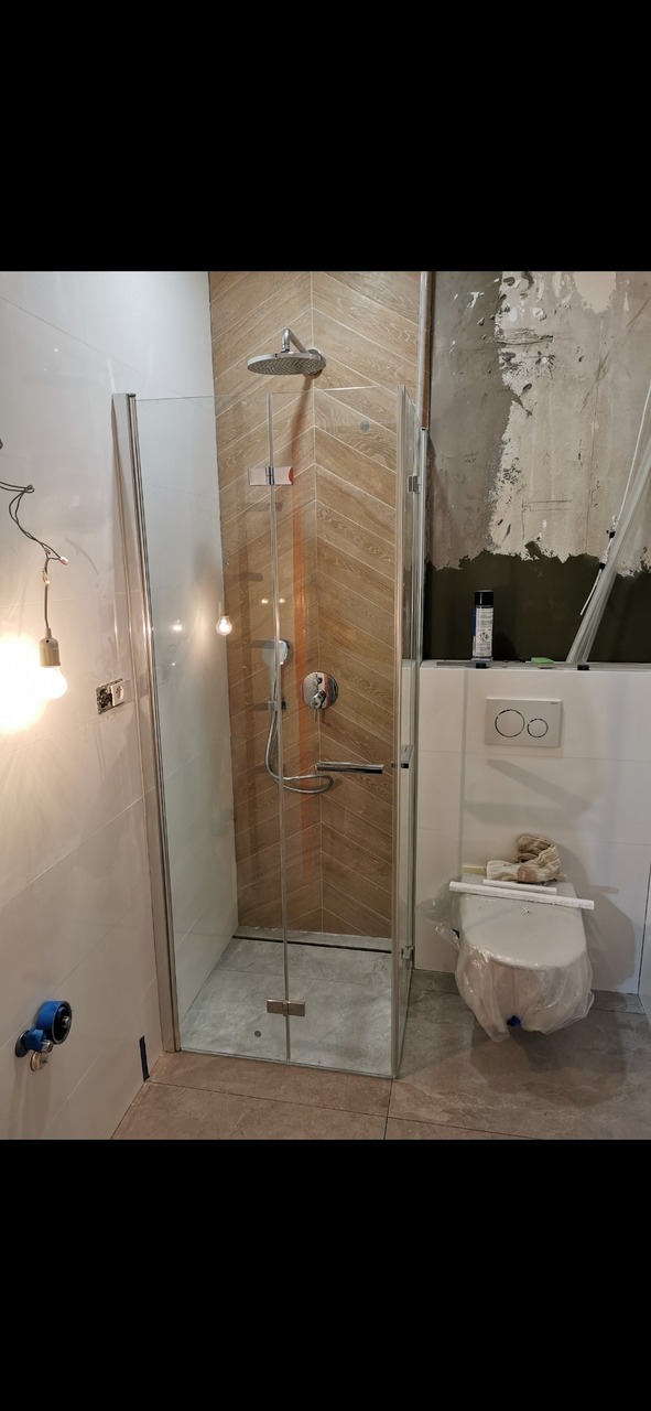 Montaż kabiny prysznicowej z deszczownicą, widoczne płytki imitujące drewno ułożone w jodełkę, częściowo wykończona ściana z widocznym tynkiem, toaleta zabezpieczona folią