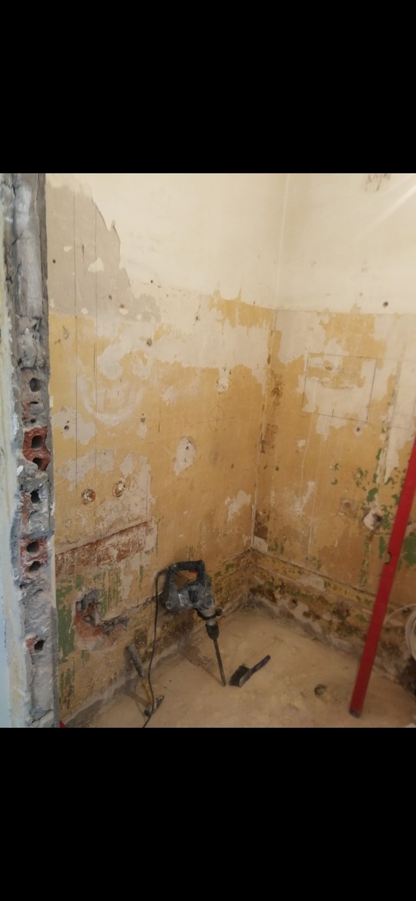 Demontaż starej okładziny w rogu pomieszczenia, widoczne ślady po instalacjach, resztki farby i zaprawy, wiertarka udarowa i szczotka na podłodze.