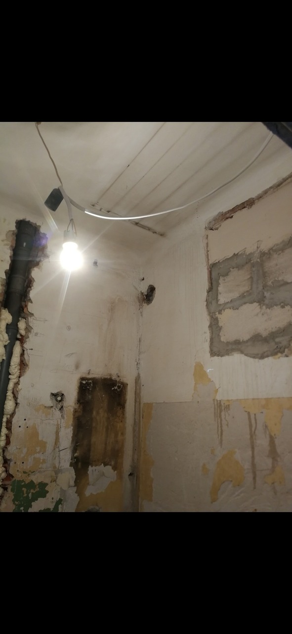 Wnętrze w trakcie remontu: odsłonięte ściany z resztkami starej farby i tapet, fragmenty muru z cegieł, rury uszczelnione pianką montażową, surowa żarówka na kablu oświetlająca pomieszczenie.