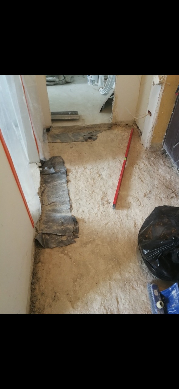 Przygotowanie podłoża pod posadzkę w trakcie remontu, widoczna poziomica, folia ochronna przy ścianie, worki na gruz i narzędzia.