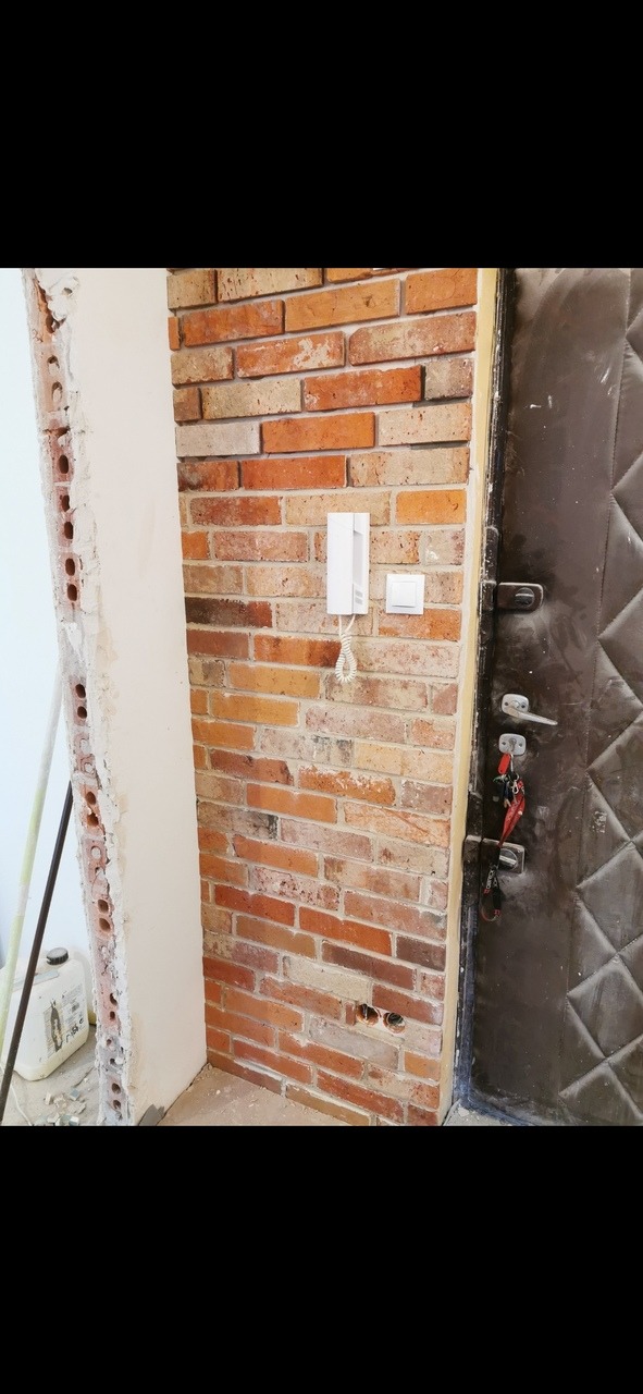 Fragment ściany z cegły klinkierowej, na której zamontowano domofon i włącznik światła, obok widoczna ościeżnica drzwi wejściowych i fragment surowej ściany przygotowanej do wykończenia.