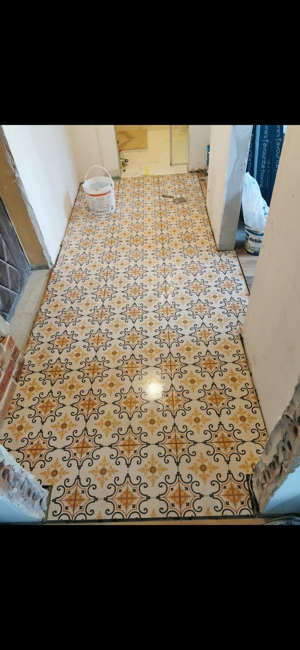 Ułożona podłoga z wzorzystych płytek w korytarzu, widoczne niedokończone prace wykończeniowe przy ścianach, wiaderko z farbą w tle.