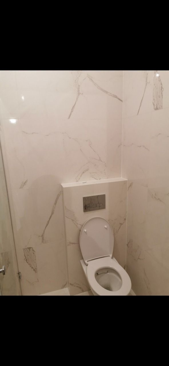 Nowoczesna toaleta z otwartą deską, obudowana płytkami imitującymi biały marmur z szarymi żyłami, w tle widoczny fragment szklanej kabiny prysznicowej.