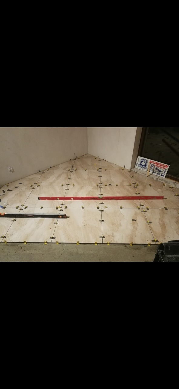 Układanie beżowych płytek podłogowych w rogu pomieszczenia, z użyciem poziomicy i klipsów poziomujących, widoczne narzędzia i naklejka reklamowa na szybie okna.