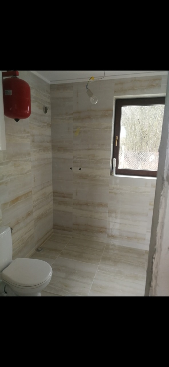 Łazienka w trakcie remontu, wyłożona płytkami imitującymi marmur, widoczny bojler, okno z widokiem na drzewa, surowa żarówka na kablu.