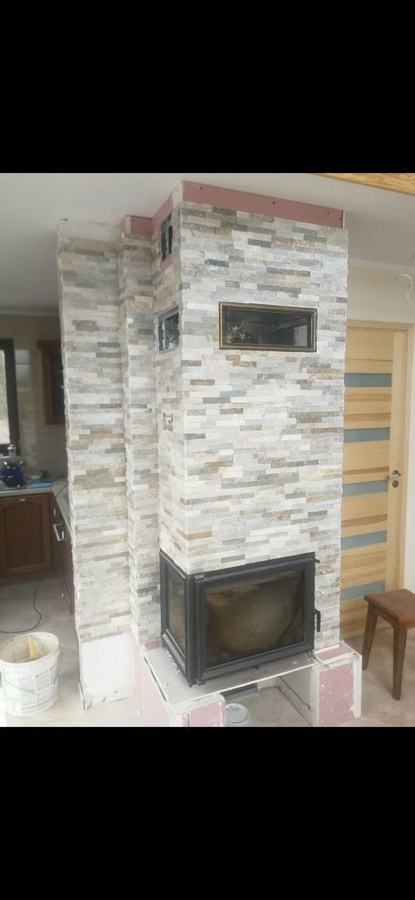 Budowa kominka z obudową z kamienia dekoracyjnego w trakcie prac wykończeniowych, widoczny wkład kominkowy z czarną ramą, fragment kuchni w tle.
