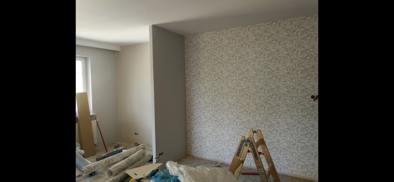 Pokój po tapetowaniu, widoczna drabina, rolki tapet i narzędzia malarskie, wzór liści na tapecie.