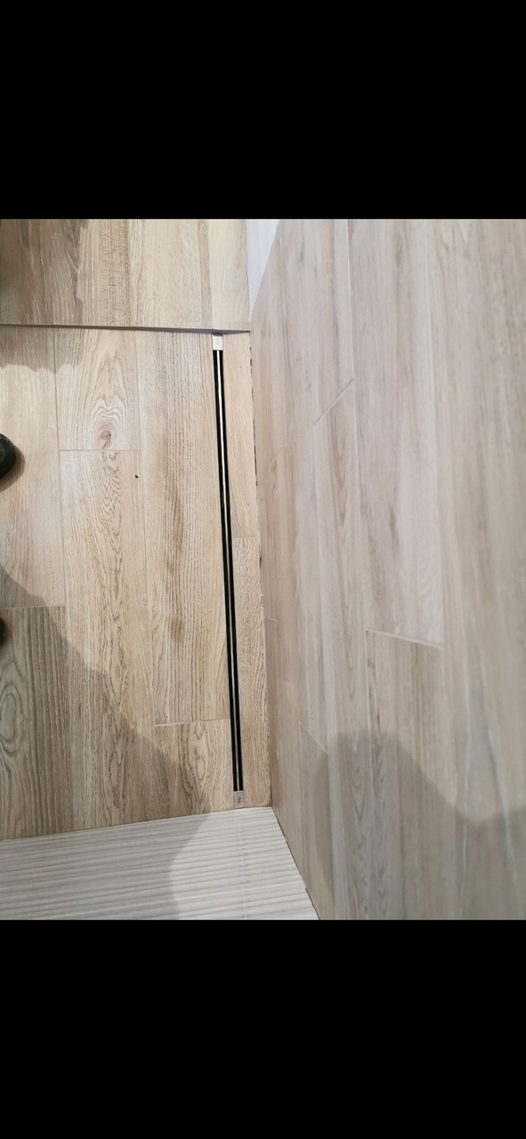 Ukończona łazienka z odpływem liniowym w podłodze wyłożonej płytkami imitującymi drewno, z widocznym fragmentem buta w lewym górnym rogu.