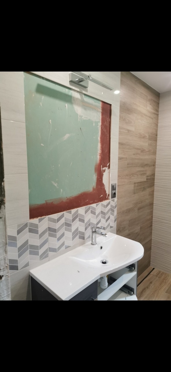 Niedokończona łazienka z umywalką i fragmentem ściany z płytek ułożonych we wzór jodełki, obok surowa ściana przygotowana pod lustro i wykończona płytkami imitującymi drewno.