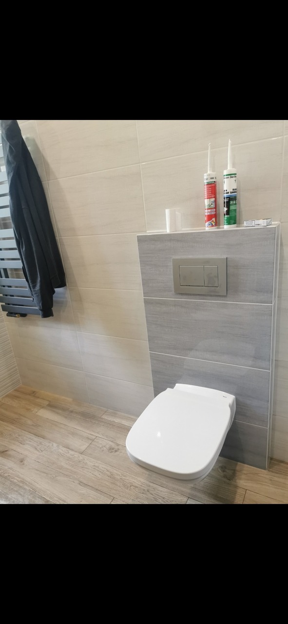 Nowoczesna toaleta podwieszana z szarym panelem spłukującym, wykończona jasnymi płytkami imitującymi drewno, z grzejnikiem drabinkowym i wiszącym ubraniem po lewej stronie, oraz tubami silikonu...